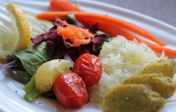 A plate of delicious ferments including sliced dill pickles, sauerkraut, fermented tomatoes, fermented garlic, fermented carrots on a bed of greens with a fresh lemon wedge. 